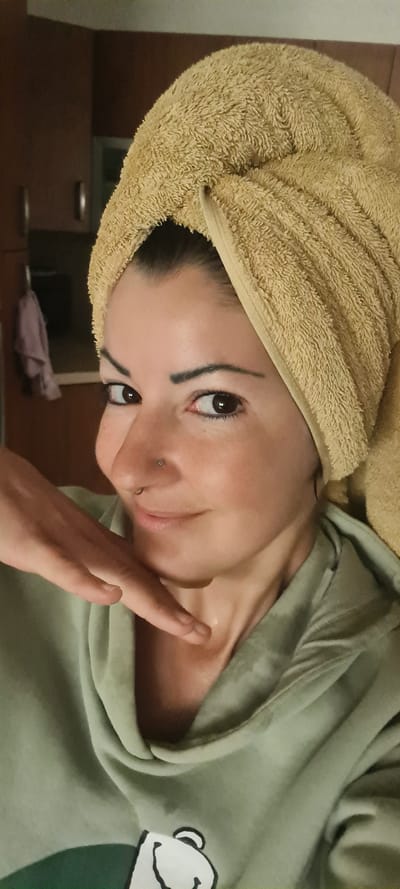 A woman with a towel wrapped around her head is applying lotion to her face. She is wearing a light green hoodie with a cartoon bear face on the right side. She has dark hair, dark eyes, and a nose piercing. The background appears to be a room with brown cabinets. The image does not reveal the location in Nahariyya, Israel.