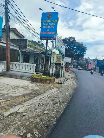 The image shows a street in Lembang, Indonesia. It's a narrow, paved road with a small, cracked concrete shoulder on the right side. A large sign advertising "Pusat Oleh-Oleh Bolu Susu Lembang & Tiramisu" is positioned at the edge of the shoulder. There are several houses and buildings visible on the left side of the road, with a white picket fence separating the road from the buildings. A few trees are visible in the background, along with a few other signs and a car parked in a driveway. Two motorbikes are visible in the distance on the right side of the road.