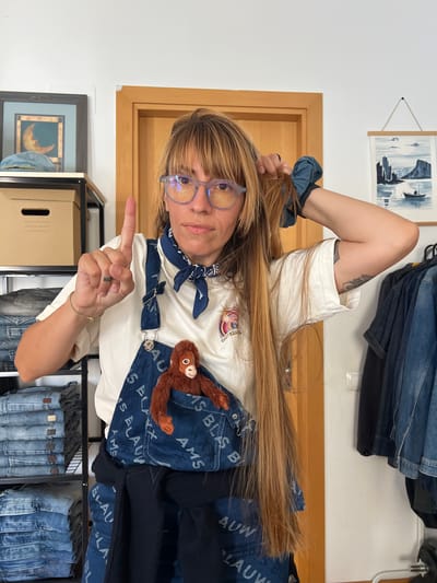 A woman with long blonde hair and glasses is standing in a room. She is wearing a white t-shirt, denim overalls, and a blue bandana. A small stuffed orangutan is peeking out of the pocket of her overalls. She is holding up her index finger on her right hand. To her left, there are shelves stacked with folded jeans and boxes. To her right, there is a wooden door and a wall with framed artwork and hanging denim clothes. The room appears to be a domestic space, possibly a bedroom or closet, located in Mollet del Vallès, Spain.