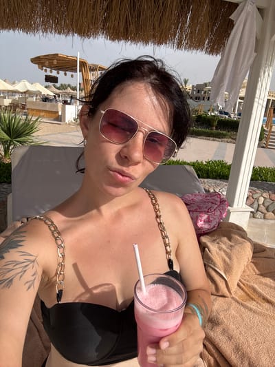 A woman in Al Ahia', Egypt, is reclining on a sunbed beneath a shaded structure, likely at a resort or beach club. She is wearing sunglasses and a black bikini top with a chain detail. She is holding a pink smoothie with a white straw. A towel is nearby. In the background, there are beach umbrellas, tables, and chairs, indicating a beachfront setting. Other buildings and landscaping are visible.