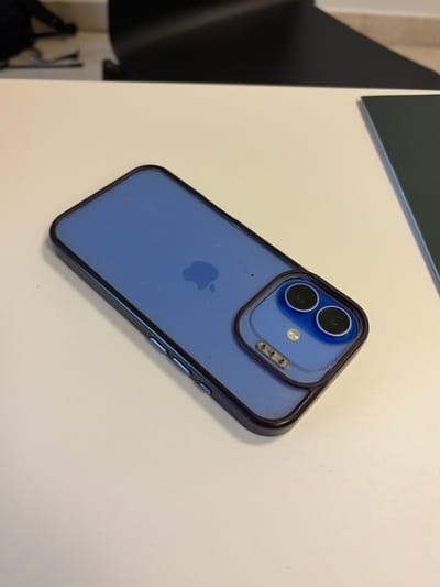 A blue Apple iPhone, likely an iPhone 12, 13, or 14 model, is positioned rear-upwards on a light-colored, flat desk surface in Alcobendas, Spain. The device is encased in a translucent protective cover featuring a dark, possibly black, perimeter and an integrated clear lens protector for its dual rear camera system. The Apple logo is visible on the phone's blue back panel through the case. Fine particulate matter and smudges are evident on the phone's surface beneath the case and on the case itself, particularly around the camera module and along the edges. Volume controls are discernible on the phone's left side. The blurred background to the upper left reveals a dark, indistinct object, while the upper right background shows a partial dark, flat rectangular item, consistent with a notebook or folder, also on the desk surface. No individuals or active interactions are present in the scene.