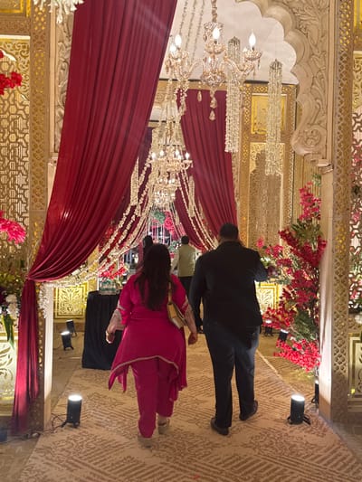 A scene depicts a decorated interior, possibly for a wedding. Red draping and crystal chandeliers adorn the ceiling. A man in a dark suit walks alongside a woman in a bright pink ensemble, each heading in the same direction. The patterned floor leads to a backdrop filled with more floral arrangements and intricate designs. Spotlights illuminate the event.
