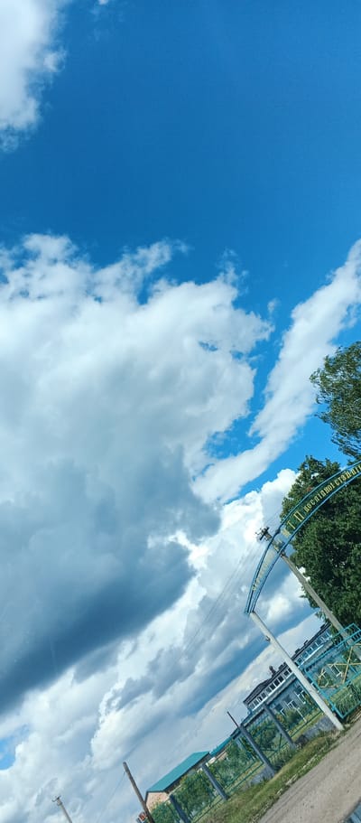 The image is a low-angle, wide shot of a sky filled with dramatic clouds, ranging from bright white cumulus to darker, more ominous storm clouds. A bright blue sky is visible in the upper portion of the frame. In the foreground, a blue arched gateway with yellow lettering is visible, partially obscured by lush green trees. The text on the gateway appears to be in Ukrainian and reads "МТП ПО ДСОПІДНОЇ СТАНЦІЇ," which translates to "MTF PO DSOPIDNE STATION." To the right of the gateway, a multi-story building with many windows can be seen, suggesting an institutional or research facility. A green metal fence runs along the property line in front of the building. Electrical poles and wires are also present in the scene, indicating utility infrastructure. The ground in the foreground is a dirt or gravel surface. The overall scene suggests an outdoor environment, possibly on the grounds of an agricultural or research station in the city of Kholodnianske, Ukraine, under a dynamic and changing sky. No people are visible in the image.