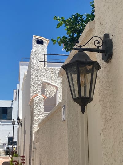 The image shows a narrow, sun-drenched street in Estepona, Spain. The architecture is predominantly white with textured stucco walls. In the foreground, a black, ornate outdoor lantern hangs on a wall. Behind it, a white building with a rough stucco finish features a prominent chimney with a tiled cap. Further back, other white buildings are visible, some with dark shutters. Green foliage cascades from above the building on the right. The sky is a clear, vibrant blue. A street lamp and plants are visible on the left side of the image. The scene suggests a warm, Mediterranean climate and a typical Spanish coastal town aesthetic.