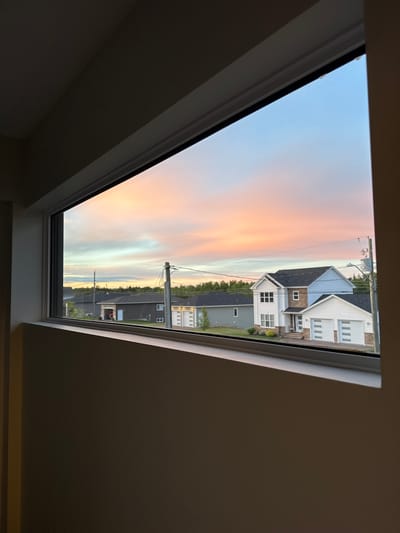 A wide, horizontal window frames a suburban street scene at dusk in Dieppe, Canada. The sky is a vibrant mix of pastel pink, orange, and blue, with wispy clouds. The foreground is dominated by the interior of a room, with neutral-colored walls and window trim. Outside, a row of modern houses with dark roofs and light-colored siding lines the street. One house in the center is particularly prominent, featuring white siding, stone accents, and a dark roof. It has multiple windows and a double garage with white doors. Power lines and poles are visible against the sky. The overall impression is one of a quiet, residential evening.