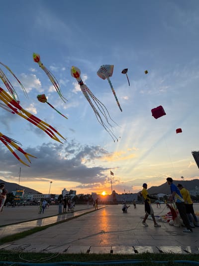This image captures a vibrant outdoor scene, likely in Quy Nhon Ward, Vietnam, during what appears to be a kite festival or a gathering where people are flying kites. The sun is low in the sky, casting a warm, golden light and creating long shadows, indicating it is either sunrise or sunset. The sky is a mix of clear blue and dramatic, wispy clouds, with visible sun rays filtering through.
The primary subjects are numerous kites of various shapes and sizes, soaring against the sky. Many are long and segmented, resembling sea creatures like octopuses or stingrays, adorned with colorful patterns. There are also simpler, square-shaped kites.
In the foreground and along a paved walkway, several groups of people are present. Some are actively engaged in flying the kites, holding onto strings, while others are observing or strolling. The people are dressed in casual attire. Some are silhouetted against the bright sky and reflecting surfaces. The setting appears to be a waterfront or a spacious open area adjacent to buildings and a mountain range in the distance. The overall mood is lively and festive, with a sense of community and enjoyment of the outdoor activity.
Notable details include the reflection of the sky and sunset on the wet-looking paved surface, suggesting recent rain or a deliberate effect. The presence of palm trees and the architectural style of some buildings hint at a tropical or subtropical coastal location. There is some visible text on signage on buildings in the background, but it is not clearly legible.