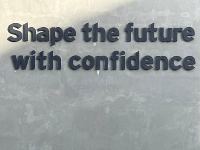The image shows the phrase "Shape the future with confidence" displayed in dark, three-dimensional lettering on a light gray concrete wall. The lighting is uneven, with shadows cast on the upper part of the text. There are slight blemishes and imperfections on the wall's surface and on some of the letters, which indicate weathering or wear. The location is Colombo, Sri Lanka. No people are visible.