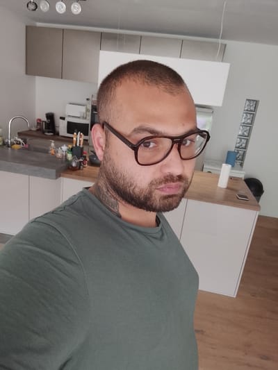 The image displays a man in his late 20s or 30s, captured in what appears to be a selfie. He has short, dark hair, a beard, and wears dark-rimmed glasses, looking directly at the camera with a neutral to slightly serious expression. A tattoo is visible on the left side of his neck. He is wearing a dark olive-green t-shirt.

The setting is a modern kitchen, likely within a home in Waasmunster, Belgium. The background reveals a clean, contemporary design with light-colored walls. To the left, a grey, stone-effect countertop features a stainless steel sink, a faucet, and various bottles and cleaning supplies. Further back, a coffee machine and a microwave are visible on a light wooden countertop, alongside kitchen utensils in a holder. Above, there are sleek, handle-less cabinets in light brown and white. To the right, another light wooden countertop holds a smartphone, a white cylindrical object, and a blue container, with a black waste bin beneath. Decorative square wall art is mounted on the far right wall. The floor is made of light-toned wood. Overhead, three recessed lights are visible on the ceiling, along with a hanging wire. The lighting suggests it is daytime indoors. No specific activity is taking place other than the man posing for the camera.