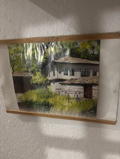 A painting is displayed on a white, textured wall. The painting depicts a traditional Bulgarian house with a stone-tiled roof and white walls, surrounded by lush greenery. The house has multiple windows, some open, and a wooden door. A smaller outbuilding with a similar roof is visible to the left. The foreground is filled with tall, green grass. The painting is bordered by two wooden strips, one at the top and one at the bottom, holding the artwork in place. The image is likely a representation of a scene in Shipkovo, Bulgaria, a village known for its traditional architecture and natural beauty.