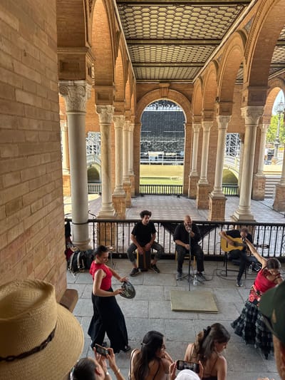 The image depicts a performance on a tiled patio in Seville, Spain, likely within the Plaza de España given the distinctive brick architecture and arched colonnades. In the foreground, the back of a straw hat worn by an observer is visible. Further back, a group of people are enjoying a live performance. A woman in a red top and black pants, holding a tambourine, is dancing with dynamic arm movements. To her left, three men are providing musical accompaniment: one plays a guitar, another sings into a microphone while seated, and a third plays a cajón. Behind the performers, a body of water is visible, crossed by a bridge, and a large, dark screen or backdrop dominates the far end of the plaza. The architectural style suggests a warm, sunny day.