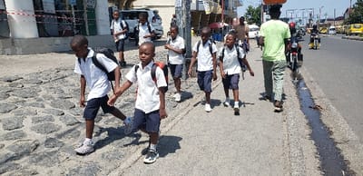The image shows a group of seven children, likely elementary school students, walking on a paved sidewalk in Rubavu, Rwanda. The sidewalk is made of stone and is located in the city center, near the intersection of a busy street and a smaller side road. The children are wearing school uniforms, consisting of white shirts and navy blue shorts. They all have black backpacks. The students are walking in a single file line, with the first child leading the group and the other six following in order. All of them are facing the same direction. 
The street is paved with asphalt and has a small patch of mud on the right side of the sidewalk, in the image’s foreground. Traffic flows on the street in both directions. There is a man walking on the sidewalk, a man riding a motorcycle on the street, and another man is crossing the street in the background. There is a parked car in the background, behind the children, and a row of houses across the street. The image was taken on a sunny day with clear skies. The children are walking toward a destination that is not visible in the frame.