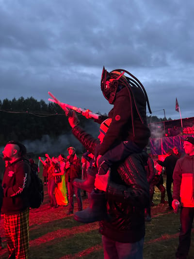 The image depicts an outdoor gathering taking place in Bolshaya Kivara, Russia, under an overcast, twilight sky. The scene is heavily illuminated by artificial red light, casting a dominant crimson hue across the foreground and midground. The ground consists of uneven dirt and sparse patches of grass.
In the center of the frame, a person wearing a dark, ribbed jacket and a distinctive helmet resembling a "Predator" alien, complete with dark dreadlock-like appendages, is perched on the shoulders of another individual. This lower individual wears a dark, possibly padded or leather jacket and a striped beanie (orange, blue, and white). Their right arm is extended upwards, holding two glowing, cylindrical objects that emit red/pink light, resembling light sticks or toy sabers. Their left arm is wrapped around the legs or waist of the person on their shoulders, seemingly for support.
To the left of the central figures, a man with dark skin and dreadlocks styled in an updo wears a dark hoodie with white lettering (partially visible as "JO...") and red and yellow plaid pants. He carries a dark backpack on his left shoulder and is looking towards the right.
To the far right, a man wearing a dark baseball cap and a two-toned jacket (dark on top, red on bottom) holds a silver beverage can in his left hand, resting at his side.
In the midground, several other individuals are visible, also bathed in the red light. Some are seen with their arms raised, possibly dancing or reacting to an event. One person in the midground is notably wearing a bright, possibly neon green or yellow garment. Wisps of white smoke or haze are present in this area, catching the red light.
In the background, a dense line of dark evergreen trees extends across the horizon. To the far right, a dark structure, possibly a stage or temporary shelter, is discernible, with a dark flag (featuring a lighter emblem) flying from a pole near it. The overall atmosphere suggests an outdoor concert, festival, or rave event.