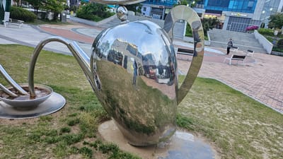 The image captures a sunny, outdoor scene in Ulsan, South Korea. A large, highly reflective, metallic teapot sculpture dominates the foreground, positioned on a patch of grass. The teapot is tilted, and its spout pours into a metal dish. The reflection on the teapot's surface mirrors the surrounding environment, including a building and a person standing on a brick pathway. Behind the teapot sculpture, a brick-paved area features white benches and a staircase leading to a building labeled "OLIVE YOUNG." A lone individual stands near the top of the stairs. The background includes trees and other buildings, indicating an urban setting.