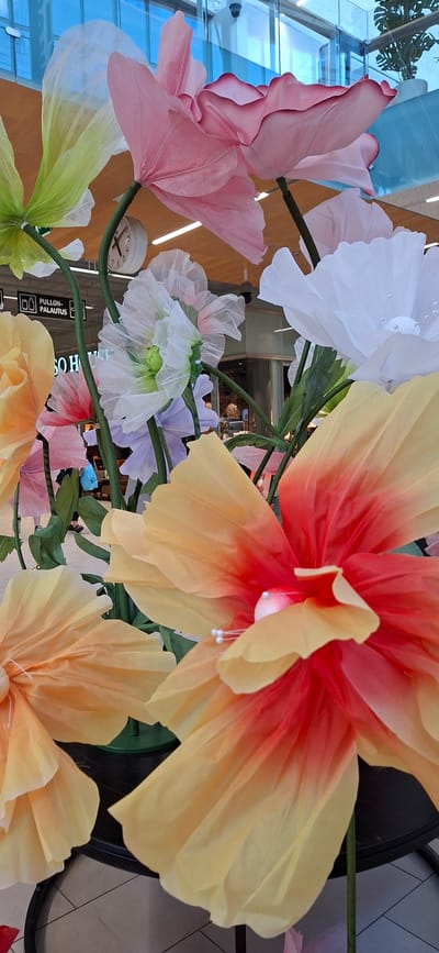 A close-up, low-angle shot showcases a vibrant display of oversized, artificial flowers in varying colors and sizes. The flowers, made from translucent material, exhibit delicate folds and gradients, with some featuring red and orange hues transitioning to yellow, while others are pink, white, and pale green. The largest flower in the foreground, predominantly yellow and red, dominates the frame with its expansive petals. In the background, blurred elements suggest an indoor setting, possibly a shopping mall or public space in Lahti, Finland, indicated by a circular clock on the wall and signage in Finnish. Reflections of a glass-covered ceiling and blue sky are visible in the upper portion of the image. The overall composition emphasizes the artificial nature and scale of the floral installation.