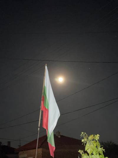 The image shows a Bulgarian flag hanging under a night sky with a bright moon. The flag is predominantly white, green, and red. Overhead wires crisscross the frame. In the upper right corner, foliage is visible near a building. The image was taken in the city of Kardam, Bulgaria.
