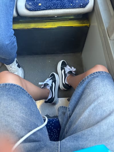 The image captures a close-up view of two people's legs and feet, dressed in denim and sneakers, sitting on what appears to be public transportation, likely a bus or train. The scene is set indoors, with the background showing a worn, grey floor and a yellow vertical pole. To the left, a blue seat with a speckled pattern is partially visible, along with a silver metallic frame.
One person is wearing black and white Vans sneakers with white laces, paired with a denim skirt or shorts. Their leg is bent, and their foot is resting on the floor. The other person, whose legs are closer to the foreground, is wearing white sneakers with black accents and white laces. They are also wearing denim, likely jeans, which are slightly frayed at the bottom. The overall lighting suggests it could be daytime, with no strong shadows indicating a specific time. The mood is casual and everyday, focusing on the footwear and clothing. No visible text is present in the image. The cropped sections provide detailed views of the sneakers and denim fabric. The location context points to Şişli, Turkey, which implies this is a scene from a public transit system in that area.