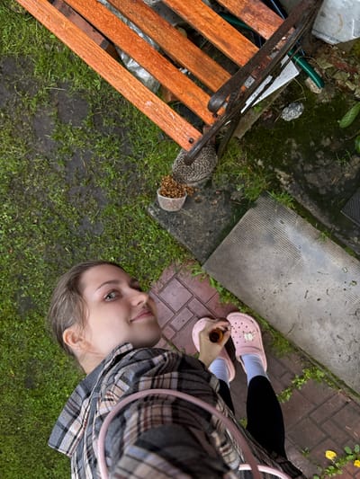 An overhead, wide-angle shot captures a young woman with fair skin and light brown hair standing in what appears to be a backyard or garden area in Ленинградская, Russia. She is looking upwards and to her right, with a slight smile. She is wearing a grey and brown plaid shirt, black leggings, and pink clogs. In her right hand, she holds a small, orange-topped object, possibly a bottle.
The ground is a mix of wet, green grass and reddish-brown brick paving. To the left of the frame, partially visible under an orange wooden slatted bench, is a hedgehog. Next to the bench, on a grey concrete slab, is a clear plastic cup filled with small, brown pellets, likely food for the hedgehog. The bench has a weathered, dark metal frame. In the background, to the right of the bench, a green garden hose is coiled. The overall environment suggests an outdoor, somewhat unkempt space.