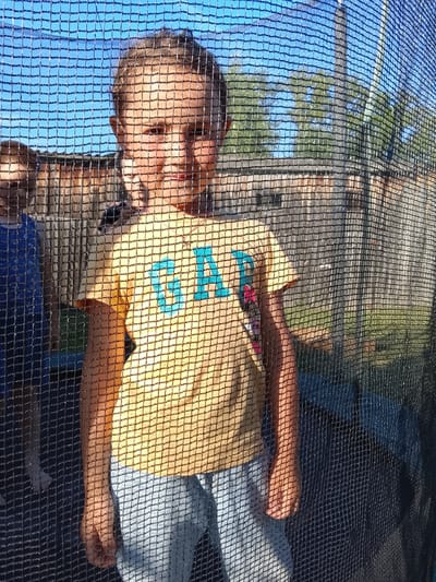 A young person is centered in the frame, viewed through a black trampoline safety net. The person, likely female, has light skin and blonde hair, and is wearing a yellow t-shirt with the letters "GAP" in blue and a red and pink design on the left chest. They are also wearing light blue jeans. Their face is partially obscured by the net, but their eyes, nose, and mouth are visible. Their right arm is bent and their hand is resting near their chest. Another person, partially visible on the left, is wearing a dark blue top. The background consists of a wooden fence, green grass, and trees under a clear blue sky. The scene appears to be a backyard in a suburban or rural setting. The image was taken in Kuyeda, Russia.