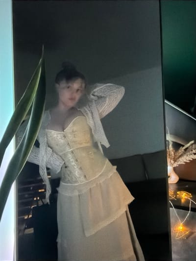 This media file captures an indoor scene, primarily featuring a television or monitor screen displaying an image or video.
**Subjects:** The main subject displayed on the screen is a young woman. She is wearing a light-colored, possibly cream or white, elaborate dress or costume. The attire includes a fitted, corset-style bodice with visible lacing or metallic stud details, and long sleeves with delicate lace patterns. Her hair appears to be styled up. Her face is slightly blurred and indistinct due to the image quality and angle.
In the immediate foreground, obscuring the bottom of the screen, is a green plant with long, pointed leaves, likely a snake plant or similar foliage.
In the background of the actual room, above the screen, various decorative elements are visible: a dark green wall or surface, a piece of dried botanical decor resembling pampas grass, a string of warm-toned fairy lights with geometric, star-shaped wire cages, and a small, white, sculpted object, possibly a decorative candle holder.
**Activity or event taking place:** The primary activity depicted is the viewing of visual media on a screen. The woman on the screen appears to be posing or captured in a still frame from a video. The image on the screen is rotated 90 degrees clockwise, showing the woman lying on her side relative to the screen's frame.
**Notable details:** The scene suggests an indoor setting, possibly an evening, given the presence of decorative fairy lights emitting a warm glow. The background behind the woman on the screen is a plain, dark grey or black, with some vertical, darker elements in the lower right, possibly indicating a staircase or structural detail within her depicted scene. The room itself, where the photo is taken, appears to be furnished with decorative items, creating a cozy atmosphere. The location context provided is Большая Шильна, Russia, referring to where the photograph was taken.
**Visible text:** There is no visible text in the image.