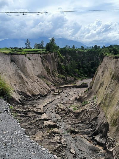 The image depicts a dramatic geological formation, likely a canyon or a deep gully, carved into the earth. The sheer, eroded walls of the gully are composed of layered sediment in shades of tan, grey, and brown, with some patches of green vegetation clinging to the slopes. The floor of the gully appears to be a dry, rocky riverbed with evidence of past water flow, showing intricate patterns of erosion and debris. On the left side of the frame, a gravelly path borders the edge of the gully. In the background, a lush green landscape with trees and hills is visible, leading to distant mountains shrouded in mist. The sky is filled with dramatic, billowing white clouds against a blue sky, suggesting a dynamic weather system. A power line stretches across the upper portion of the image, supported by unseen poles. The scene is a natural landscape, likely in a rural or semi-rural area, showcasing the erosive power of nature. The provided location information places this scene in Kebunbaru, Indonesia, suggesting a tropical or subtropical environment given the vegetation. There are no people or animals visible in the image, and no specific actions or interactions are depicted.