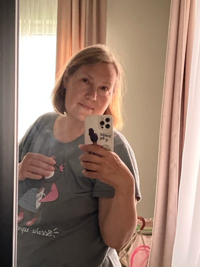 A woman is taking a mirror selfie in a room. She is wearing a dark gray t-shirt with a cartoon character on it. She is holding a smartphone in her right hand, and her left hand is on her chest, near the cartoon. The background shows curtains and a window. The lighting suggests it is daytime. The scene is an indoor residential setting.