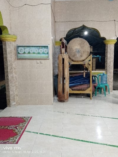The image shows the interior of a building in Kecamatan Kroya, Indonesia, located at latitude -6.423 and longitude 107.996. There is a large wooden drum resting on a wooden frame. The drum is made of brown wood and has a round shape. The drum is resting on a wooden frame that is also made of brown wood. The frame has several shelves, and there are some folded blue fabric on the shelves. In the background there is a clock on the wall. The floor is tiled and there is a red rug on the floor. The walls are tiled and painted white. The ceiling is white. In the right corner of the image, there is a green plastic chair. The image was taken on 18 April 2025 at 18:01.