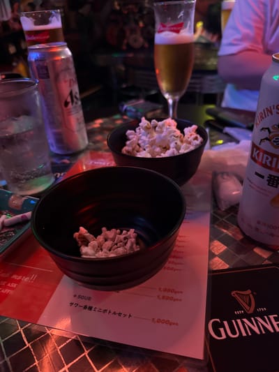 This is a medium shot, taken at an angle, of a table in what appears to be a dimly lit bar or restaurant in Adachi, Japan. The dominant subjects are two black bowls filled with popcorn, placed on a surface with a checkered pattern. To the left of the bowls, a glass of beer with a red logo, possibly Budweiser, is visible. Behind it, a white can with "KIRIN'S" and "KIRI" written on it is also present. The table also features a Guinness coaster with the iconic harp logo and text "ESTD 1759 GUINNESS". A menu is laid out on the table, displaying prices in yen, with items like "SOUR" and "サワー各種ミニボトルセット" (various sour mini bottle set) listed. The lighting suggests it's evening or nighttime. The overall mood is casual and relaxed, possibly a night out with friends. There is no visible human presence, but the scene implies people are either about to eat or have just finished, with the drinks and popcorn suggesting a leisurely activity. The text is clear and legible, offering specific details about the beverages and prices.