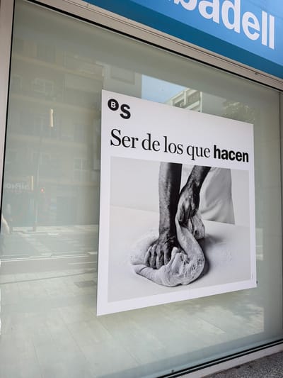 This is a close-up shot of a poster displayed behind a glass window, likely a shop window or a building facade, in Ibiza, Spain. The poster is printed in black and white and features a dramatic close-up image of two hands intertwined and covered in what appears to be flour or a similar powdery substance. One hand seems older, with wrinkled skin, while the other is smoother. The image conveys a sense of texture and detail, suggesting an activity involving dough or pottery.

To the left of the image, a portion of a blue sign with white lettering is visible, displaying the word "adell". The poster itself has bold black text running vertically along its left side. The text reads "BS Ser de los que hacen". The poster is angled, with the top edge closer to the viewer and extending towards the right.

The setting appears to be outdoors, as evidenced by the reflection of buildings and what looks like shadows of trees on the glass in the background. The lighting suggests it might be daytime, possibly sunny, given the clarity of the reflections and the contrast in the image. The overall mood of the poster, with the hands and the text, could evoke themes of creation, craftsmanship, or community.