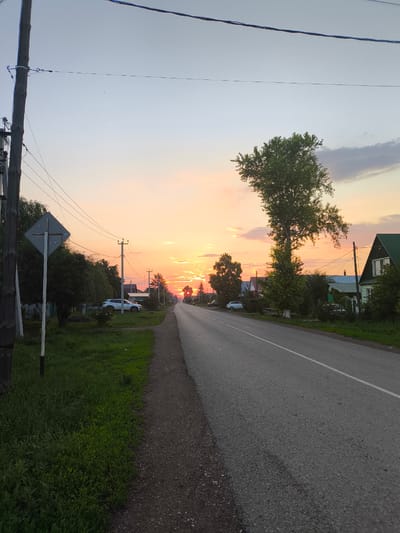 The image depicts a rural road at sunset in Saraktash, Russia. A paved road runs from the foreground into the distance, with a dirt shoulder on the left and a grassy verge. Power lines crisscross the sky, supported by wooden poles. Several houses with pitched roofs, some painted green, line the right side of the road, interspersed with trees and fences. A large, leafy tree stands prominently on the right. On the left, a diamond-shaped road sign is visible next to a wooden utility pole. The sky is filled with the warm hues of sunset, with streaks of orange and pink clouds. No people are visible in the scene. The overall impression is one of a quiet, semi-urban or rural setting as day transitions into evening.