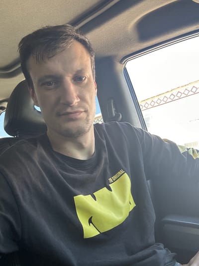 A person is taking a selfie inside a vehicle. The person is a white male with short, dark brown hair and a light beard and mustache. He is wearing a black long-sleeved shirt with a large, bright yellow graphic on it. The graphic appears to be a stylized smiley face. Text above the graphic reads "THE ORIGINAL". The vehicle appears to have a sunroof open, revealing a bright sky, suggesting daytime. The interior of the car is visible, including a dark headrest. In the background outside the vehicle, a building with a diamond-patterned facade is visible. The image seems to have been taken in Makiv, Ukraine. The person's expression is neutral.