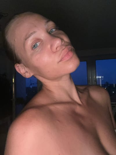 The media file is a close-up, vertically oriented selfie of a woman with fair skin and blue eyes. She is looking directly at the camera with a neutral expression. Her head is tilted to her left, and her gaze is slightly downward.

She is positioned in the foreground, with a dark, out-of-focus background that appears to be an interior space with a window. Through the window, a night sky is visible, with a hint of city lights or buildings in the distance, suggesting an urban setting. The lighting is dim, casting soft shadows on her face and shoulder.

The woman's upper body is visible, including her shoulders and neck. She is not wearing any clothing from the visible area. There is a small earring in her left ear.

The context provided is "Mühldorf a. Inn, Germany," which might indicate the geographical location where the photo was taken. The time of day is clearly night, due to the dark sky and artificial lighting cues. There are no discernible objects or text in the image. The activity is a self-portrait, likely taken by the subject herself.
