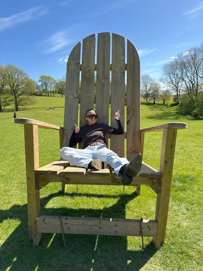 A medium shot, taken outdoors, features a woman lounging on an oversized wooden Adirondack chair set amidst a lush green lawn, against a backdrop of trees and a clear blue sky with wispy clouds, suggesting a rural setting.
The woman, clad in a dark long-sleeved shirt, light-colored jeans, and black boots, wears sunglasses and reclines casually in the chair, giving a thumbs-up gesture. Her positioning and expression suggest a relaxed and playful mood.
The Adirondack chair itself is constructed from unfinished wood. The expansive lawn, typical of a park or large garden, is well-maintained, and distant trees add depth to the scene.
While the exact location within the city of Sevenoaks, United Kingdom, is impossible to pinpoint, the presence of a large, grassy area suggests a park or expansive garden.