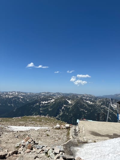A mountain landscape under a clear blue sky. The foreground features a rocky, uneven terrain with patches of snow and scattered stones. A concrete structure, possibly part of a building or viewing platform, is visible on the right side, with some snow accumulating on its surface. In the middle ground, a range of mountains stretches across the horizon, characterized by jagged peaks, dark green vegetation, and patches of white snow. The sky is a vibrant blue with a few wispy clouds scattered across it. The image appears to be taken from a high vantage point, offering a panoramic view of the mountainous region. The location is likely the Rila Mountains, near Samokov, Bulgaria, possibly at the terminus of a ski lift or a mountain hiking trail. There are no people or animals present in the image.
