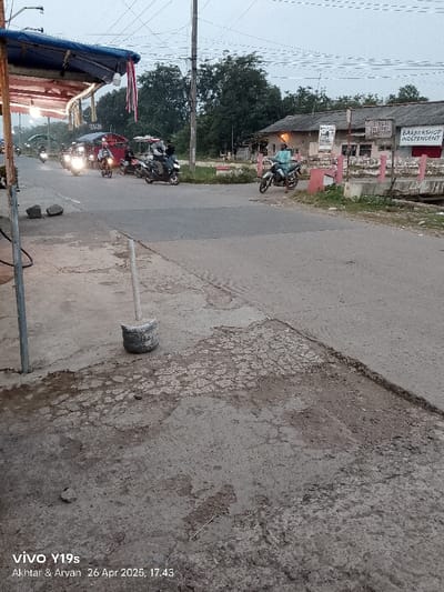 The image shows a street in Kecamatan Kroya, Indonesia, with a cracked and broken concrete surface, a grey cement block, and a metal post. The street is lined with trees and buildings, including a barber shop with a sign that says “Barber Shop Independent”. Several motorcycles are parked in the distance, and one motorcycle is parked on the side of the road with a man sitting on it. There is a red and pink metal railing to the right of the scene and a sign on a building to the left. The sky is overcast and there is a red and white cloth hanging from a wooden structure with an awning in the foreground. The image was taken with a Vivo Y19s phone. The timestamp is 26 Apr 2023, 17:43. The location coordinates are -6.422 latitude and 107.996 longitude.