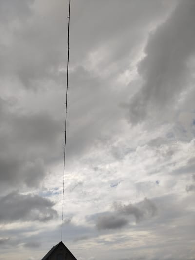 The image depicts a cloudy sky over a rooftop in Крывасельскі сельскі Савет, Belarus. A thin, dark line, likely a wire or antenna, extends vertically from the peak of the rooftop upwards into the sky, marked by several small, equally spaced attachments. The sky is filled with a variety of clouds, ranging from dark, heavy formations to lighter, more dispersed ones, with hints of blue sky visible in patches. The rooftop is triangular, with a dark surface and a lighter-colored shuttered window. The overall atmosphere suggests overcast or impending weather conditions.