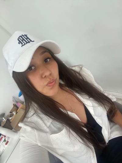 A young woman, wearing a white baseball cap with a dark emblem and a white button-down shirt over a black top, poses for a selfie in what appears to be an indoor setting with a plain white wall as the background. She has long, dark hair and is looking directly at the camera with her lips pursed. A gold necklace with a pendant is visible on her chest. To the left of the frame, a shelf unit with various items, including what look like art supplies or stationery, can be seen. The lighting suggests it is daytime. The location is not specified in the image, and therefore it cannot be stated to be in Akyar, Russia.