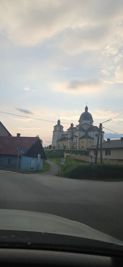 The image shows a view of the historic Church of the Assumption of the Blessed Virgin Mary in Niebylec, Poland, at dusk. The church, a prominent yellow and white structure with multiple domes and spires topped with crosses, is situated on a hill. In the foreground, a narrow paved road curves uphill towards the church. To the left of the road is a small, weathered blue wooden building with a red-tiled roof and a pedestrian crossing sign. Further to the right, a beige house with a dark roof is visible, and beyond it, the church complex. The sky is filled with soft, pale clouds, suggesting the end of the day. The overall scene depicts a tranquil, late afternoon or early evening atmosphere in a small Polish town.