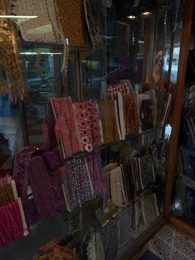 The image presents a close-up, slightly angled view into a multi-tiered glass display cabinet located within a retail establishment in Santander, Spain.
The cabinet, featuring dark wooden framing on the right side, is extensively stocked with various textile trimmings, ribbons, and decorative laces. These items are systematically arranged on at least five visible glass shelves. The top shelf displays gold-toned glittered fabric and white lace. The middle shelves exhibit an array of pink, fuchsia, and red ribbons, some with white floral appliques, alongside gold metallic braids, brown velvet trims, and various other textured and patterned ribbons. The lower shelves showcase purple embroidered ribbons, vivid purple sequined trims, fuchsia flat ribbons, and a selection of cream, beige, and darker patterned laces, including leopard print and geometric designs.
Prominent reflections are visible on the cabinet's glass panels. A person with long, light-colored hair, presumably the photographer, is reflected on the upper-right section of the cabinet. Further reflections suggest external elements, including what appears to be a light-colored vehicle and the general store interior.
The floor in the lower right portion of the image displays blue and white patterned tiles, arranged in a geometric design. No direct human action or interaction is depicted, beyond the implied presence of the reflected photographer.