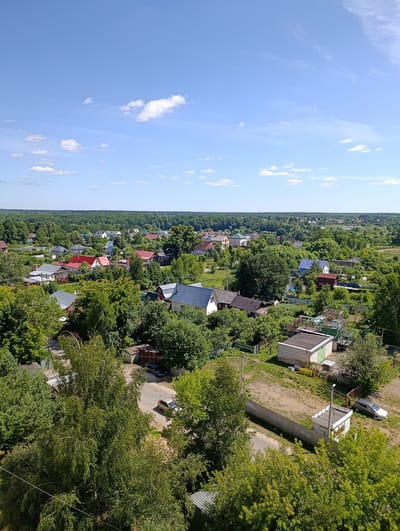 This is a high-angle, daylight, full shot of a semi-rural residential area in Alexandrov, Russia. The scene is dominated by lush green trees and vegetation interspersed with numerous houses with various roof colors, including red, gray, and blue. A few roads are visible, one of which has a silver car parked on it and another with a light-colored car driving away. Several small outbuildings and fences are also present. The sky is a clear blue with scattered white clouds. The overall impression is of a tranquil, well-treed community under a bright sky.