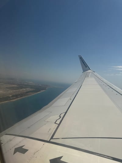 This media file is a photograph taken from an airplane window, looking down at the coastline. The primary subjects are the wing of the airplane and the landscape below. The wing, a large, white structure with black accents and rivets, occupies a significant portion of the frame. The text "RYANAIR" is clearly visible on the wingtip.
The scene below depicts a coastal area with a body of water and land. The land shows signs of development, with buildings, roads, and possibly an airport or industrial area. The colors are muted, with the blue of the sky and water transitioning into a hazy, brownish hue over the land, suggesting the time of day might be late afternoon or early morning, or perhaps some atmospheric pollution. There are some faint clouds visible in the upper sky. The overall impression is of a commercial flight in progress, with the photographer observing the world from a high altitude. There are no people visible. The photograph is oriented vertically, as if taken while tilted.
Notable details include the "RYANAIR" logo on the wing. The landscape below provides a hint of the location. The presence of a large structure resembling an airport and the proximity to the sea are key visual cues. Some text on the wing reads "NO STEP". The weather appears to be clear, though the haze over the land might indicate some atmospheric conditions. The emotion conveyed is one of observation and travel.