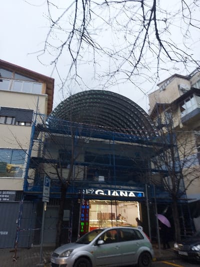 This vertical-format photograph depicts a muted, overcast urban street scene in Tiranë, Albania, characterized by buildings and bare tree branches, suggesting a late autumn or winter day.
In the mid-ground, a multi-story commercial building is prominent, featuring a large, arched structure made of corrugated metal, possibly an awning or part of its facade, supported by blue metal scaffolding or framework. A brightly illuminated sign on this building reads "GIANA" in white capital letters on a dark background. Below "GIANA," other vertically stacked, illuminated text is partially visible, appearing to spell out a word like "FASHION" or "MODISH."
To the right of this building, a silver or light grey compact car is parked on the street. Further down the sidewalk, behind the car, a bright pink umbrella is visible, hinting at cool or rainy weather. A white parking sign with blue symbols and text, possibly in Albanian, is mounted on a pole near the car.
On the left side of the frame, part of another building with a light-colored facade and a sloped brown roofline is seen. Bare tree branches crisscross the upper portion of the image, silhouetted against the grey sky and the buildings. The artificial illumination of the shop signs suggests it is either dusk or a very dark, cloudy day. No individuals are clearly visible, but the scene captures the static environment of an urban street.