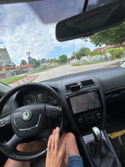 The image shows the interior of a car from the driver's perspective, looking out onto a street in Silistra, Bulgaria. A person's right hand rests on the steering wheel, which features the "SKODA" logo and an "AIRBAG" label. The dashboard includes an infotainment screen displaying a navigation map with Cyrillic text, partially showing "улица" (street) and street names like "ул. София" and "ул. Комсомолска," with a blue marker indicating the current location. A small black device, possibly a GPS or dashcam, labeled "NAVITEL," is mounted near the central air vents. The gear stick indicates a 6-speed manual transmission and is marked "4x4." A yellow banana peel is discarded on the floor near the gear shift.
Outside the vehicle, it is daytime with a bright, partly cloudy sky. The car appears to be stopped or moving slowly on a road. Visible traffic signs include a blue circular sign indicating a bicycle path, a red triangular sign (likely a yield or priority road sign), and a yellow square sign with a black arrow signifying a U-turn. Orange and white road markers separate the vehicle's lane from an adjacent path. The surroundings consist of trees, residential buildings with varying architectural styles, some featuring traditional tile roofs, and a long wall with painted murals. The scene depicts a typical urban or semi-urban street environment.