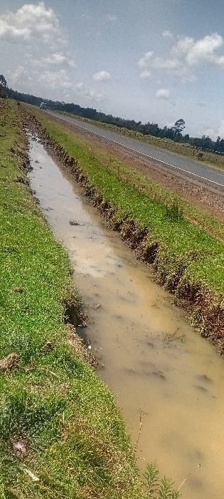 This image depicts a roadside environment located in the city of Heni Village, Kenya, under a sky with scattered white clouds and clear blue patches. A prominent, muddy water-filled drainage ditch occupies the left foreground, running diagonally towards the mid-right. The turbid, brown water within the ditch contains sparse vegetative debris. Its banks are covered with green grass. Immediately adjacent to the right of the ditch is a paved, two-lane asphalt road featuring a broken white center line, extending into the background. A light-colored vehicle is discernible in the distant portion of the road. Beyond the road, a dense line of green trees and foliage establishes the horizon.