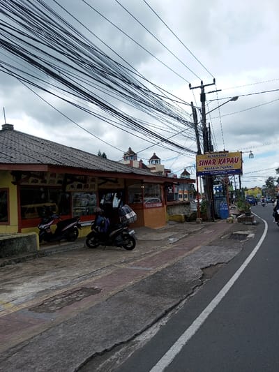 The image depicts a street scene in Taohom, Laos, under an overcast sky. A densely packed network of black utility cables spans the upper portion of the frame, originating from a tall utility pole located on the right side of the image. This pole also supports a street light and a large, yellow rectangular sign. The sign, prominently displayed, advertises "RUMAH MAKAN SINAR KAPAU" and "MASAKAN PADANG," with additional text and a phone number below.
To the left, a single-story commercial building with yellow and orange exterior walls and a grey corrugated roof extends horizontally. Its facade features multiple glass-fronted display areas. A red sign on the building's front includes the text "MASAKAN PADANG" and "MENU KATERING." Two motorcycles are parked on the concrete sidewalk directly in front of this building: a pink scooter on the left and a dark-colored scooter equipped with a large square delivery box on its rear rack to the right.
The sidewalk, constructed from concrete and paved sections, shows signs of wear and repair, including visible cracks and patches. A low concrete barrier separates the building's immediate frontage from the main sidewalk. A blue trash receptacle is positioned near the base of the utility pole.
On the right, an asphalt road curves slightly to the right, marked by a solid white line separating the shoulder or a parking lane from the main thoroughfare. Several vehicles, including motorcycles and a blue car, are visible in the distance on the road, traveling away from the viewer. Behind the commercial building, other multi-story structures are visible in the background, featuring distinct domed or cupola-shaped roofs. Green foliage is also present further in the background. No clearly discernible individuals are present in the foreground.