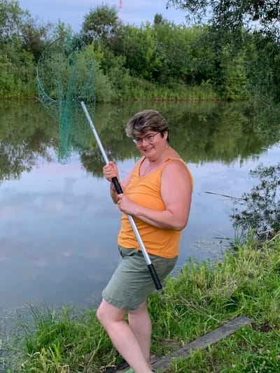 A woman stands by a body of water holding a fishing net. She is wearing an orange tank top and green shorts. The water is calm and reflects the sky and trees. The scene appears to be a natural setting with lush vegetation surrounding the water's edge. The location is described as Суроватиха, Russia.