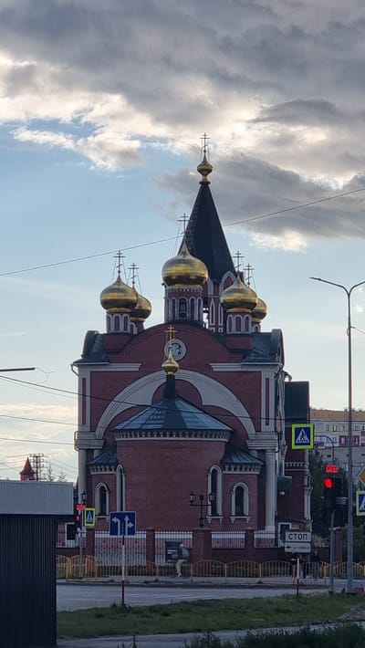 The image shows the Transfiguration Cathedral in Megion, Russia, an Eastern Orthodox church with multiple golden domes and a dark spire topped with a cross. The cathedral is primarily red brick with white architectural details. In the foreground, there is a street with traffic signs, including a pedestrian crossing sign and a traffic light displaying red. Power lines are visible overhead. The sky is partly cloudy with a mix of blue sky and gray clouds. A few people are visible walking on the sidewalk near the church.