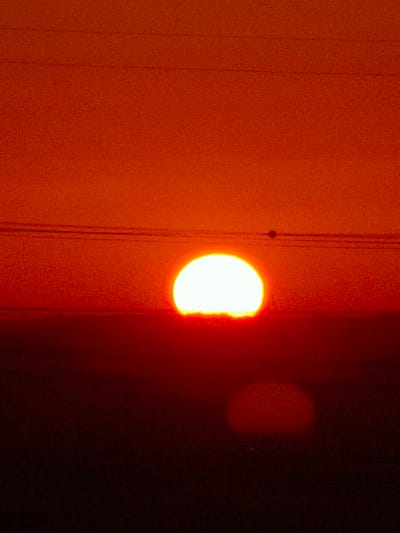 The image captures a sunset over Be'er-Sheva, Israel. A bright, white sun, appearing as a semicircle, is positioned low on the horizon, partially obscured by a dark silhouette that suggests the ground or a distant landscape. Two thin, dark horizontal lines, likely power lines, stretch across the upper portion of the sky, with one line passing just above the sun. A small, dark circular object, possibly a bird or an insect, is visible in the sky between the power lines and the sun. The sky is a gradient of deep orange and red, transitioning to a darker red and black towards the bottom of the frame, indicative of the fading daylight. The overall scene is dominated by warm, intense colors associated with a sunset.