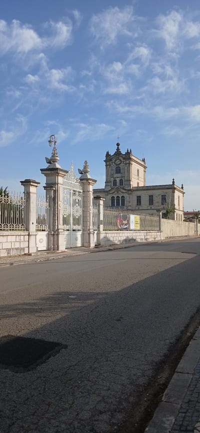 The image shows a street view of the Palace of Justice of Figueira da Foz, Portugal. The ornate, white iron gates of the palace are visible, along with a portion of the building itself. A section of paved road is also shown. The sky is blue with streaky clouds. No people are visible.