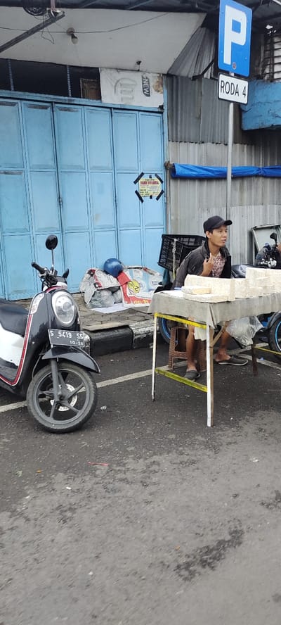 The image shows a street scene in Mojokerto, Indonesia, located at coordinates -7.467 latitude and 112.438 longitude. A man in a black baseball cap and a pink and black patterned shirt sits at a table, his legs crossed, holding a white object in his left hand. The table is covered in a grey cloth and has a stack of white objects on it. There is a black and white scooter parked on the left side of the image. The scooter has a license plate that reads "S 5419 SO 10-26". There is a blue sign with a white letter "P" and the words "RODA 4" in white on it. There is a blue tarp on the building in the background and a blue corrugated metal wall behind it. The man is sitting on a yellow stool, and there is a yellow-painted curb separating the street from the sidewalk. The ground in the image is a grey, paved road, and there is a black curb in the foreground. The building in the background is made of concrete and has a blue paint job. There are several wires hanging from the building's ceiling, and there is a sign that reads "NEW" in the background. The image is taken during the daytime, and there is no sunlight shining directly on the scene.