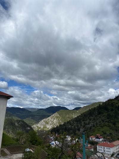The image shows a view of mountains and a town, likely Giovren, Bulgaria, based on the context. The sky is dominated by heavy, grey clouds. Below the clouds, a mountainous landscape stretches into the distance. The mountains are covered in varied vegetation, from dense forest to lighter-colored, possibly rocky, terrain. Adjacent to the mountains, rooftops and buildings of a town are visible, including buildings with red-tiled roofs and white walls. A teal-colored utility pole is in the mid-ground. In the foreground, a section of a building with a red roof is visible, suggesting the viewpoint is from an elevated position. The image orientation is rotated clockwise.
